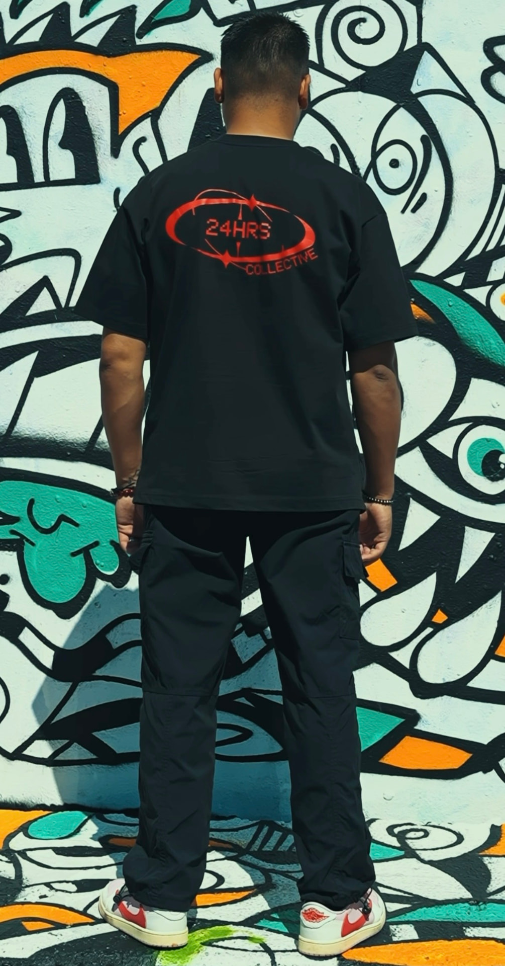 24HRS Collective tee with red logo print, worn in front of a graffiti wall
